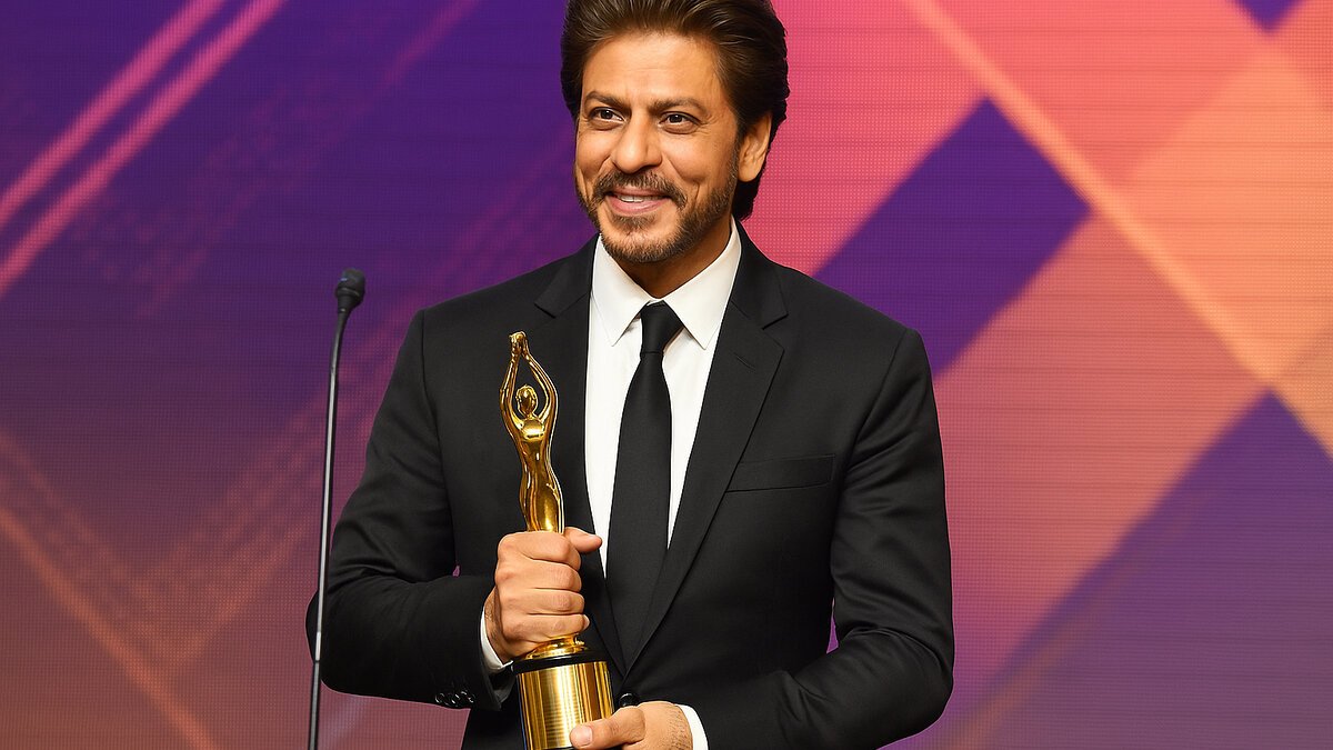 Shah Rukh Khan smiling while holding a golden trophy at an award ceremony, dressed in a black suit and tie with a colorful stage backdrop.